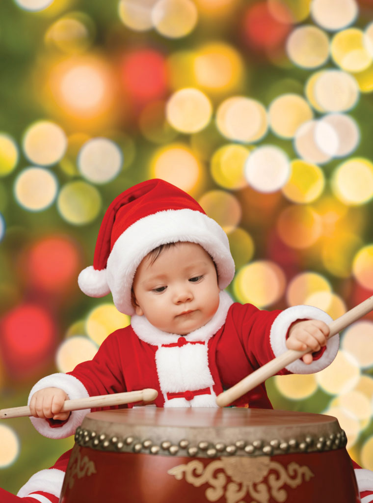 Christmas lights background with a toddler in a red santa outfit playing the drums